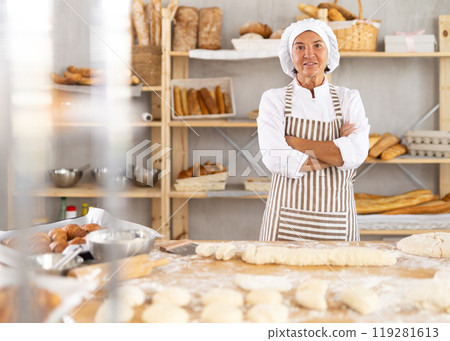 Confident senior female bakery in apron posing with arms crossed indoors in bakery kitchen Confident senior female bakery in apron posing with arms crossed indoors in bakery kitchen 119281613
