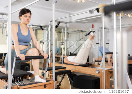Focused woman performing set of pilates exercises on reformer in fitness studio. Healthy active lifestyle concept Focused woman performing set of pilates exercises on reformer in fitness studio. Healthy active lifestyle concept 119281637