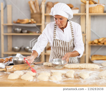 Aged woman baker buttering pieces of dough in bakery Aged woman baker buttering pieces of dough in bakery 119281638