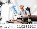 A technician operates a lathe machine in an industrial workspace, meticulously adjusting a metal 119281913