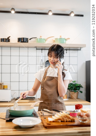 A charming Asian woman is talking on the phone while cooking in the kitchen. A charming Asian woman is talking on the phone while cooking in the kitchen. 119282034