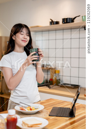 A beautiful, happy young Asian woman enjoying her drink at a kitchen island in the kitchen. A beautiful, happy young Asian woman enjoying her drink at a kitchen island in the kitchen. 119282069
