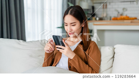 Smiling pretty young Asian woman sitting on cozy couch Smiling pretty young Asian woman sitting on cozy couch 119283664