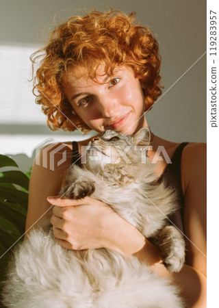 portrait red-haired curly young woman with pet gray fluffy cat in arms, lit by bright sun 119283957