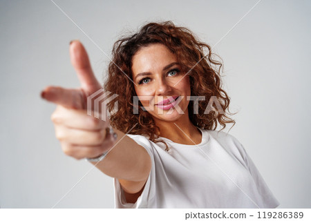 A young woman with curly hair playfully points towards the camera in a bright studio A young woman with curly hair playfully points towards the camera in a bright studio 119286389
