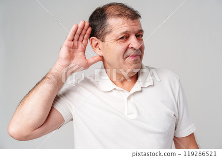 Elderly man smiling, playfully holding his hand to his ear while listening intently against a light background Elderly man smiling, playfully holding his hand to his ear while listening intently against a light background 119286521