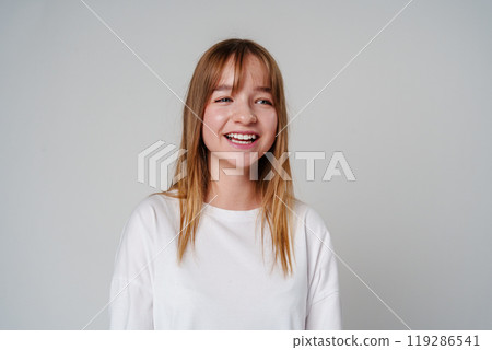 A young woman with long hair smiles brightly while wearing a simple white shirt against a neutral background A young woman with long hair smiles brightly while wearing a simple white shirt against a neutral background 119286541