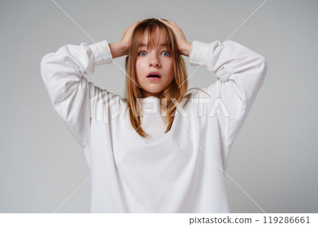 A teen girl in a loose white sweater looks surprised with her hands on her head against a plain gray backdrop A teen girl in a loose white sweater looks surprised with her hands on her head against a plain gray backdrop 119286661