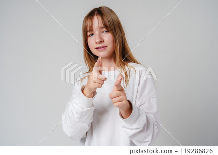 A young girl in a white sweatshirt playfully poses with finger guns against a neutral background A young girl in a white sweatshirt playfully poses with finger guns against a neutral background 119286826