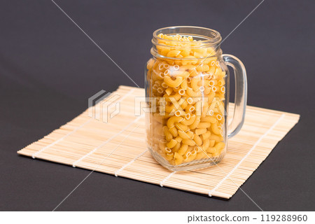 Uncooked Chifferi Rigati Pasta in Glass Jar on Bamboo Mat on Black Background 119288960
