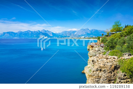Rocky seashore near Konyaalti beach, yacht with a white sail in blue Gulf of Antalya, mountains on horizon near popular resort city Antalya, Turkey 119289138