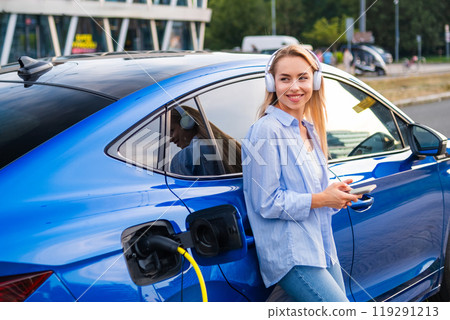 Happy blonde woman with long hair enjoys music with headphones while charging her electric vehicle at a city station 119291213