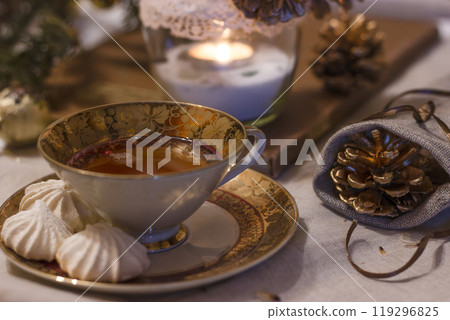 Winter still life. Cup of tea with meringues on a cozy table with a linen tablecloth on it. Beside a candle and decorative pine cones in a bag. Cozy atmosphere. Porcelain cup with gilding and painting 119296825