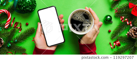Above view of female hand holding smart phone with hot cup of coffee. Christmas decoration on colored background 119297207