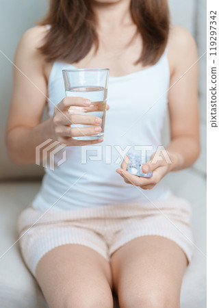 woman hand holding medicine painkiller pill and water glass on the sofa at home, taking for headaches, stomach ache, Diarrhea Pain from food poisoning, Endometriosis, Hysterectomy and Menstrual 119297342