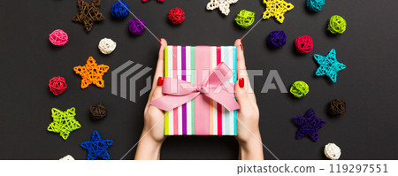 Top view of female hands holding a Christmas present on festive black background. Holiday balls and stars. New Year holiday concept 119297551