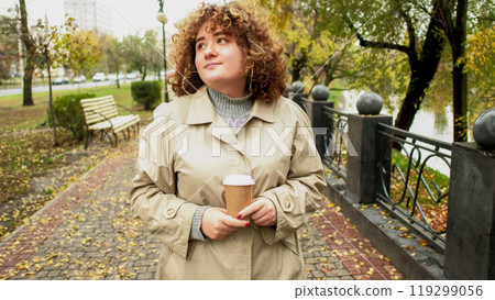 Park walk. Beautiful day. Happy peaceful smiling chubby girl enjoying fall weather strolling outside with coffee cardboard cup. Park walk. Beautiful day. Happy peaceful smiling chubby girl enjoying fall weather strolling outside with coffee cardboard cup. 119299056