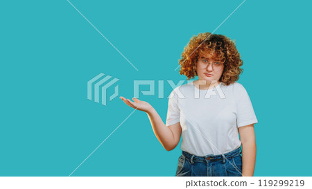 Skeptic reaction. Bored expression. Annoyed smart girl in glasses showing empty space with open palm isolated on blue empty space background. Skeptic reaction. Bored expression. Annoyed smart girl in glasses showing empty space with open palm isolated on blue empty space background. 119299219