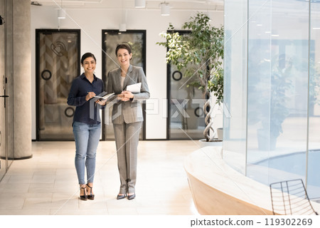 Two diverse happy confident employee women standing in office hallway Two diverse happy confident employee women standing in office hallway 119302269