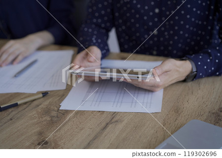 Accountants or auditors reviewing financial documents while sitting at the wooden desk in office. Professionals analyzing paper reports and using calculator and tablet computer, close up. Audit and Accountants or auditors reviewing financial documents while sitting at the wooden desk in office. Professionals analyzing paper reports and using calculator and tablet computer, close up. Audit and 119302696
