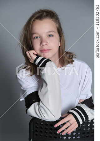 A close-up of a young girl with a calm expression, resting her chin on her hand. The casual outfit and varsity jacket create a relaxed and authentic atmosphere, perfect for youth-related themes. 119302763