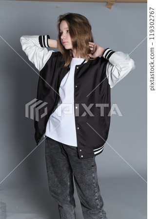 Side view of a teenage girl casually draping a varsity jacket over her shoulder. Her soft smile and laid-back posture capture a sense of youthful confidence and comfort. Side view of a teenage girl casually draping a varsity jacket over her shoulder. Her soft smile and laid-back posture capture a sense of youthful confidence and comfort. 119302767