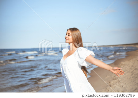 Happy smiling beautiful woman is on the ocean beach in a white summer dress, open arms 119302799