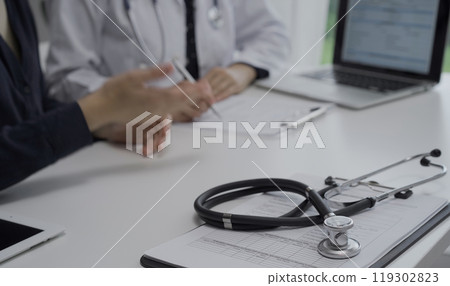 Stethoscope over a clipboard with medical papers is lying on the table near doctor and patient discussing health while sitting in clinic. Medicine and science Stethoscope over a clipboard with medical papers is lying on the table near doctor and patient discussing health while sitting in clinic. Medicine and science 119302823