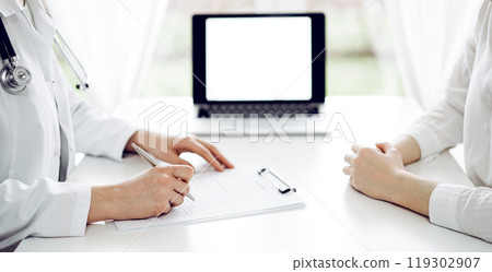 Doctor and patient sitting near each other at the white desk in clinic. Female physician is listening filling up a records form. Medicine concept Doctor and patient sitting near each other at the white desk in clinic. Female physician is listening filling up a records form. Medicine concept 119302907