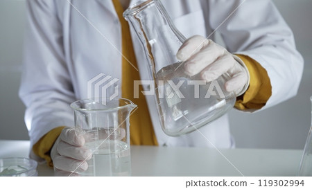 Unknown scientist wearing a lab coat and gloves is carefully pouring a liquid from one beaker into another, close up. Science and medicine concepts 119302994