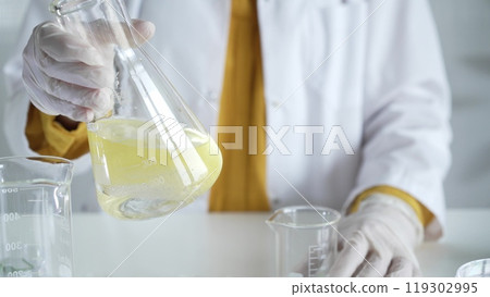 Unknown scientist wearing white protective gloves is carefully shaking up a yellow chemical solution inside an Erlenmeyer flask in a laboratory, close up. Science and medicine Unknown scientist wearing white protective gloves is carefully shaking up a yellow chemical solution inside an Erlenmeyer flask in a laboratory, close up. Science and medicine 119302995