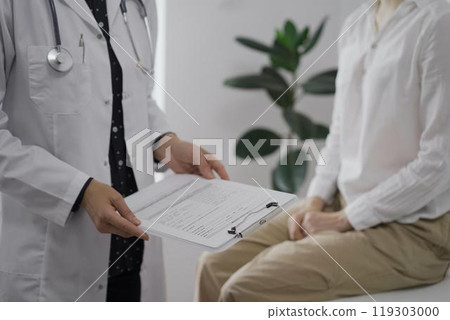 Doctor and a patient discussing something. The female physician, wearing a white medical coat is keeping clipboard with a medical notes besides a young woman during a consultation in the clinic Doctor and a patient discussing something. The female physician, wearing a white medical coat is keeping clipboard with a medical notes besides a young woman during a consultation in the clinic 119303000