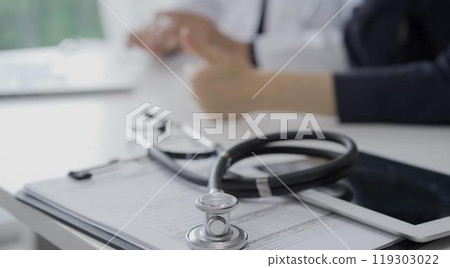 Stethoscope over a clipboard with medical papers is lying on the table near doctor and patient discussing health while sitting in clinic. Medicine and science Stethoscope over a clipboard with medical papers is lying on the table near doctor and patient discussing health while sitting in clinic. Medicine and science 119303022