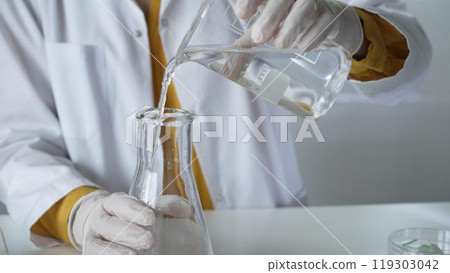 Unknown scientist with a lab coat and white protective gloves is carefully pouring a liquid from a beaker into an Erlenmeyer flask, close up. Science and medicine concept Unknown scientist with a lab coat and white protective gloves is carefully pouring a liquid from a beaker into an Erlenmeyer flask, close up. Science and medicine concept 119303042