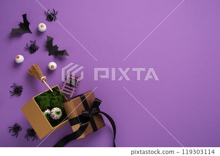Halloween gift box with spooky decorations: fake eyeballs, bats, spiders, and broomstick on purple background. Space for your text, announcement, invitation. 119303114
