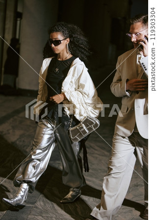 Vertical shot of glamorous Middle Eastern female celebrity wearing trendy metallic trousers paired with silver purse, confidently walking in camera lights caught together with man in city street 119303304