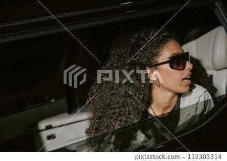 Shot through opened car window of glamorous Middle Eastern female celebrity in futuristic sunglasses caught unawares by paparazzi sitting in back seat of limousine, camera flash, copy space Shot through opened car window of glamorous Middle Eastern female celebrity in futuristic sunglasses caught unawares by paparazzi sitting in back seat of limousine, camera flash, copy space 119303314