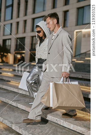 Vertical shot of fashionable male celebrity wearing classy gray suit walking with woman in trendy outfit down stairs snapped by paparazzi coming out of department store after shopping, camera flash 119303325