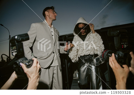 Low angle shot of glamorous female Middle Eastern celebrity pulling up hood for privacy caught by paparazzi stepping out of car, accompanied by well dressed man in suit at night street in city 119303328