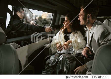 Shot from inside of luxury car of smiling Middle Eastern female celebrity interviewed by reporter through open window while sitting in back seat of car with male partner, copy space 119303388