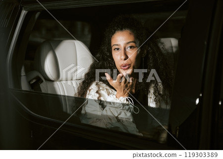 Portrait shot through open limousine window of Middle Eastern female celebrity looking at paparazzi playfully answering public attention with blowing kiss while leaving ceremony, camera flash Portrait shot through open limousine window of Middle Eastern female celebrity looking at paparazzi playfully answering public attention with blowing kiss while leaving ceremony, camera flash 119303389