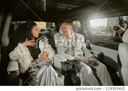 Medium shot of smiling male celebrity in classy suit talking to young Middle Eastern girlfriend sitting in back seat of car, while enjoying paparazzis attention living in public eye Medium shot of smiling male celebrity in classy suit talking to young Middle Eastern girlfriend sitting in back seat of car, while enjoying paparazzis attention living in public eye 119303402