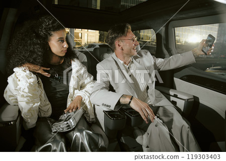 Medium shot of smiling male fashion icon in classy suit sitting in back seat of car with fashionable woman, while taking selfie with paparazzi in background 119303403