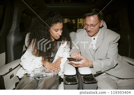 Medium shot of male celebrity in grey suit using phone looking at screen with curly Middle Eastern girlfriend sitting in back seat of car, camera flash Medium shot of male celebrity in grey suit using phone looking at screen with curly Middle Eastern girlfriend sitting in back seat of car, camera flash 119303404