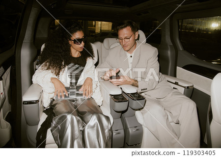 Successful male celebrity in classy gray suit using phone showing screen to fashionable Middle Eastern girlfriend sitting inside car with contemporary interior, camera flash Successful male celebrity in classy gray suit using phone showing screen to fashionable Middle Eastern girlfriend sitting inside car with contemporary interior, camera flash 119303405