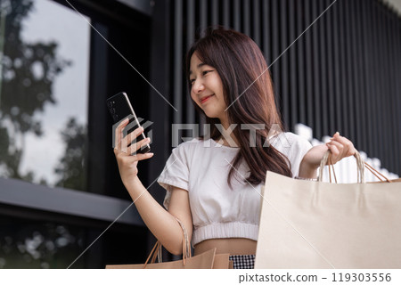 Smiling Woman Holding Shopping Bags at the Mall While Checking Her Smartphone for the Latest Deals and Offers Smiling Woman Holding Shopping Bags at the Mall While Checking Her Smartphone for the Latest Deals and Offers 119303556