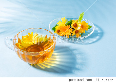 Glass cup and saucer with calendula tea and fresh flowers on light blue closeup. Light and shadows. Glass cup and saucer with calendula tea and fresh flowers on light blue closeup. Light and shadows. 119303576