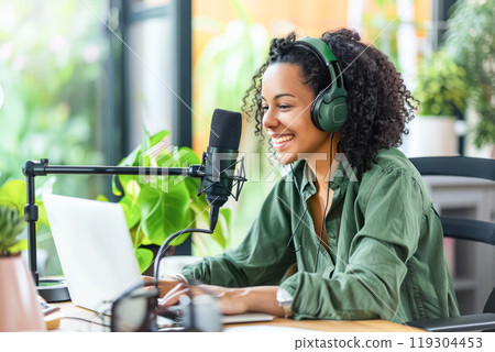Smiling woman in green shirt with voluminous hair, podcasting at home with laptop and microphone. Modern online communication, blogging Smiling woman in green shirt with voluminous hair, podcasting at home with laptop and microphone. Modern online communication, blogging 119304453