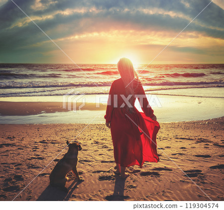 Young happy woman in a long red dress on the beach in the evening. The woman and dog gaze magical sunset together 119304574