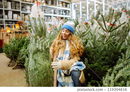 young smiling woman chooses Christmas tree to buy. Happy woman buying New Year tree for Christmas. young smiling woman chooses Christmas tree to buy. Happy woman buying New Year tree for Christmas. 119304616